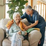 Older woman sits on a chair holding a mug while a caregiver in scrubs drapes a blanket over her shoulders in a bright living room.