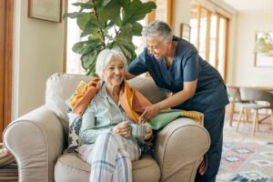 Older woman sits on a chair holding a mug while a caregiver in scrubs drapes a blanket over her shoulders in a bright living room.