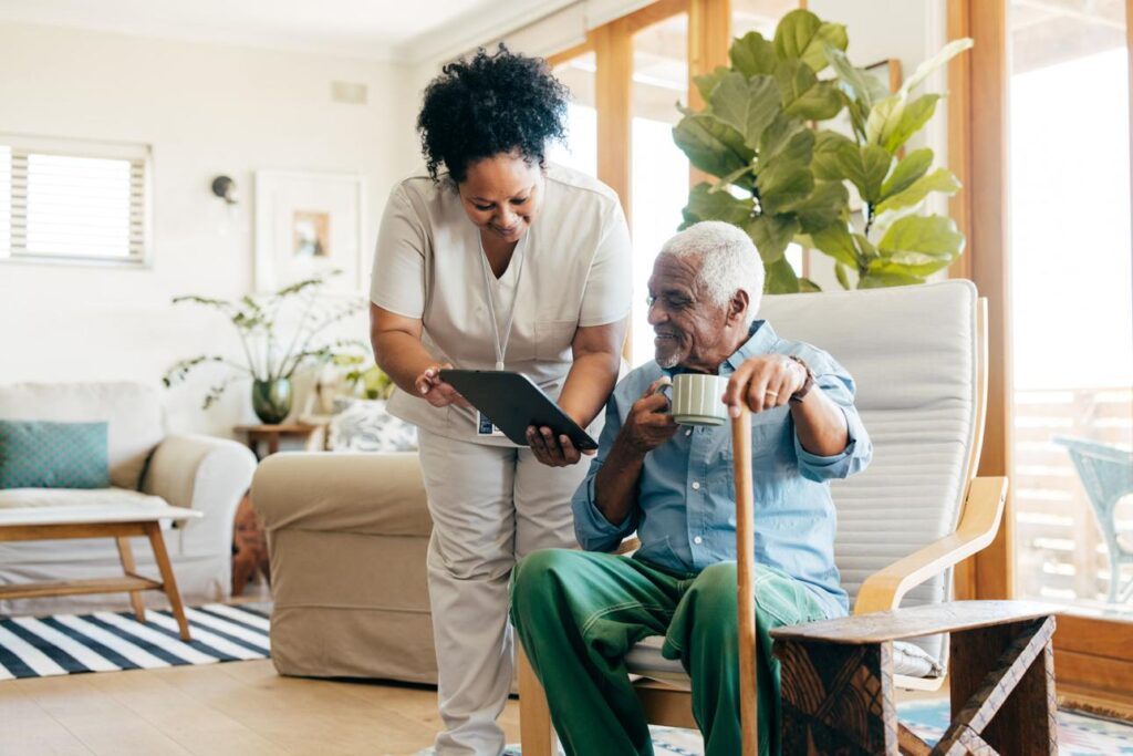 A nurse shows a tablet to an elderly man sitting in a chair, holding a cup and cane, in a bright living room.