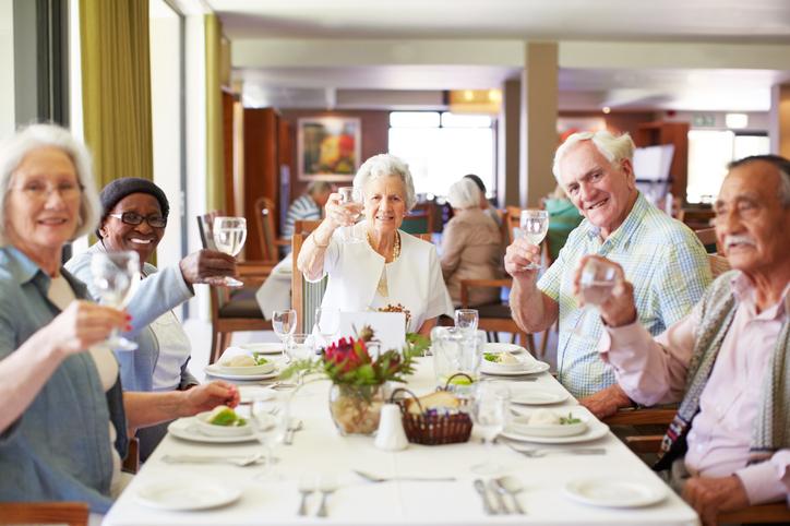 A group of five elderly people sit at a dining table in a bright room, smiling and raising glasses for a toast.