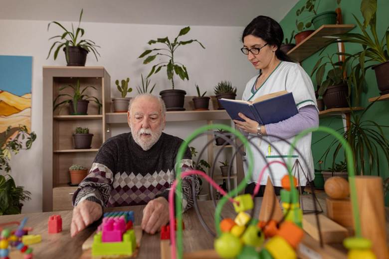 An older man arranges colorful blocks at a table while a woman in scrubs stands nearby, holding a notebook in a room with several potted plants.