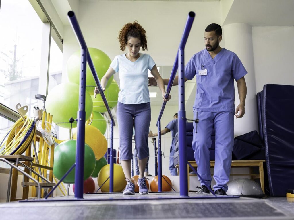A woman walks between parallel bars in a physical therapy clinic as a therapist in scrubs observes her progress.