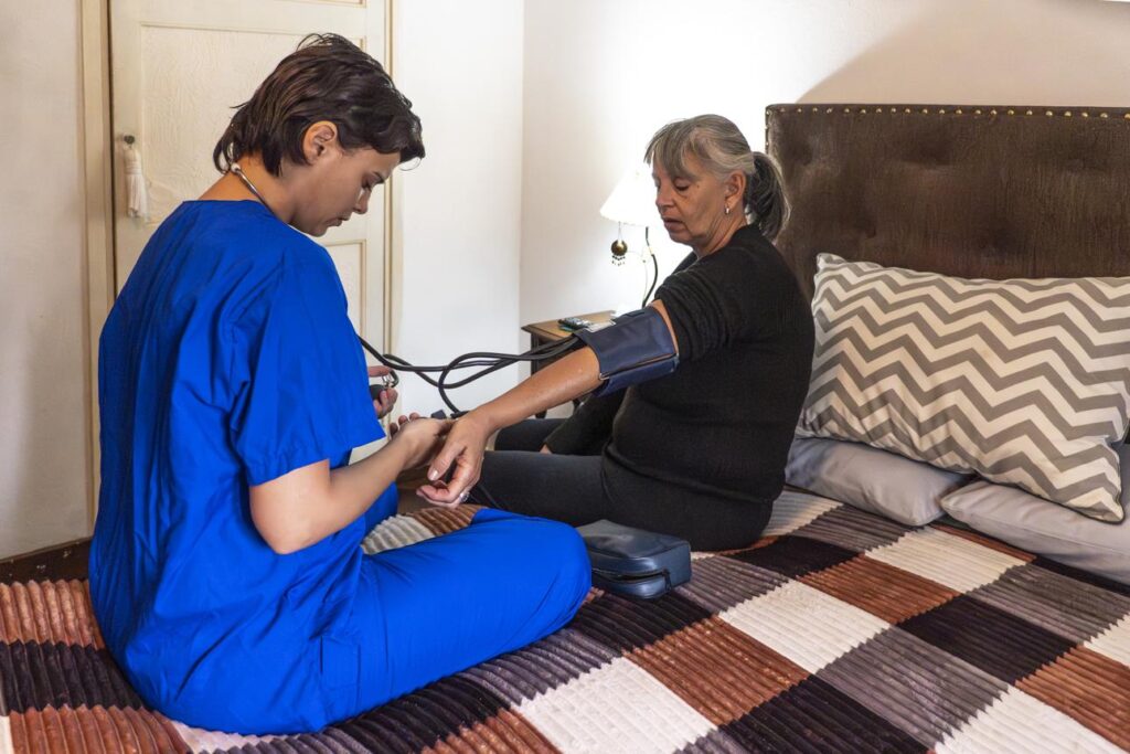 A healthcare worker in blue scrubs checks the blood pressure of an older woman seated on a bed in a home setting.