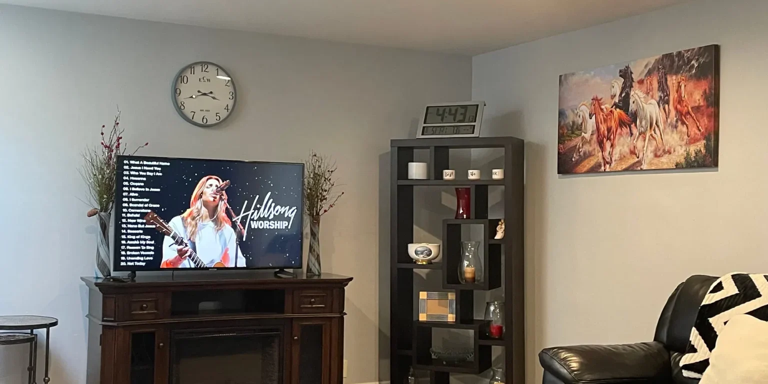 A living room with a TV displaying Hillsong Worship, a wall clock, a black shelf with decor items, a horse painting, leather furniture, and wood flooring.