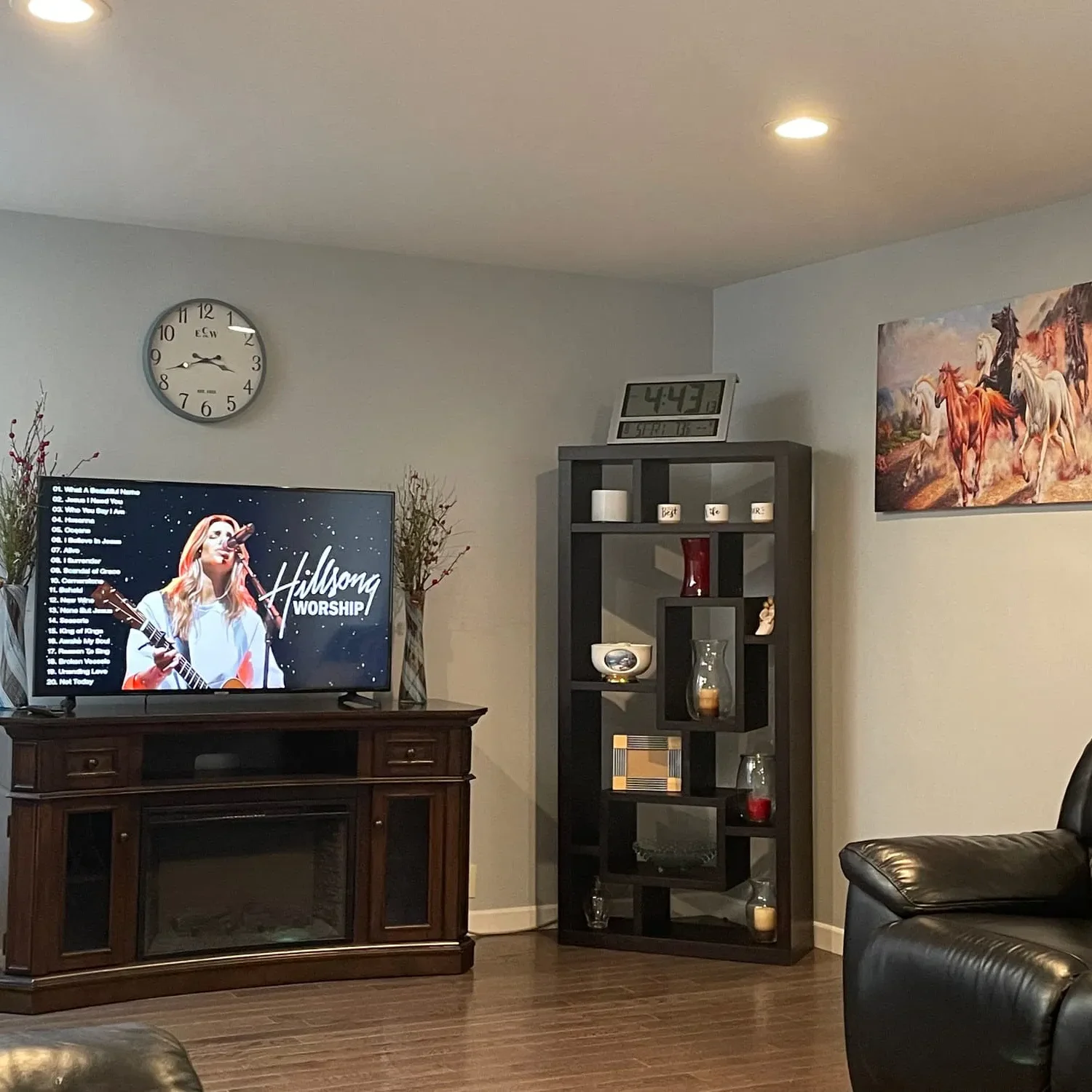 A living room with a TV displaying Hillsong Worship, a wall clock, a black shelf with decor items, a horse painting, leather furniture, and wood flooring.