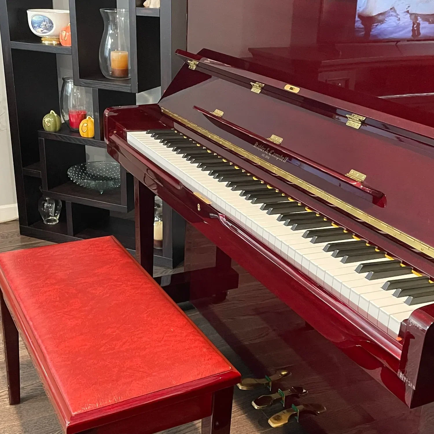 A polished red upright piano with a matching bench sits next to a black shelving unit adorned with decorative items, creating a warm and inviting atmosphere reminiscent of Everett and Arlington adult family homes.