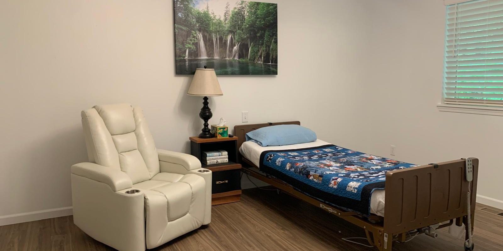 A simple bedroom in one of the Snohomish County adult family homes, featuring a hospital bed with a blue quilt, a white recliner, nightstand with lamp, and wall art of a forest waterfall.
