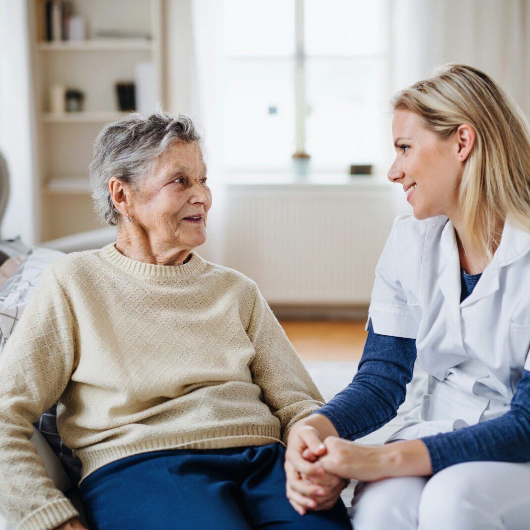 A nurse sits on a bed holding hands with an elderly woman, both smiling and engaged in conversation in a well-lit room at one of the welcoming Snohomish County adult family homes.