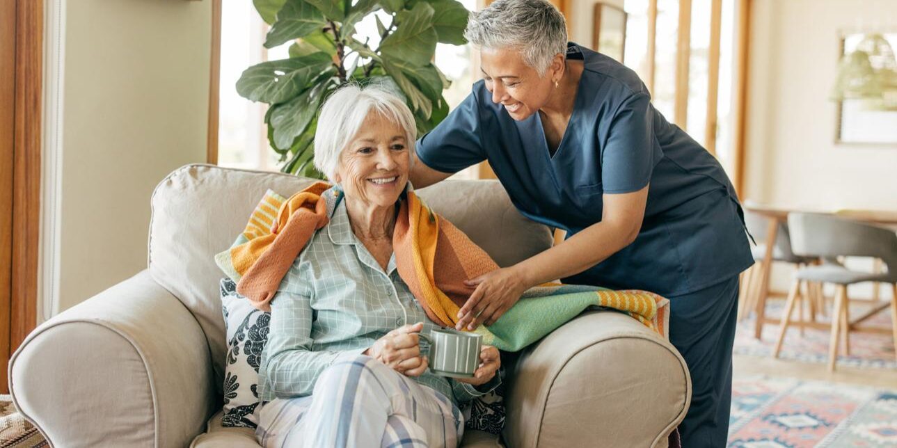 Older woman sits on a chair holding a mug while a caregiver in scrubs drapes a blanket over her shoulders in a bright living room.